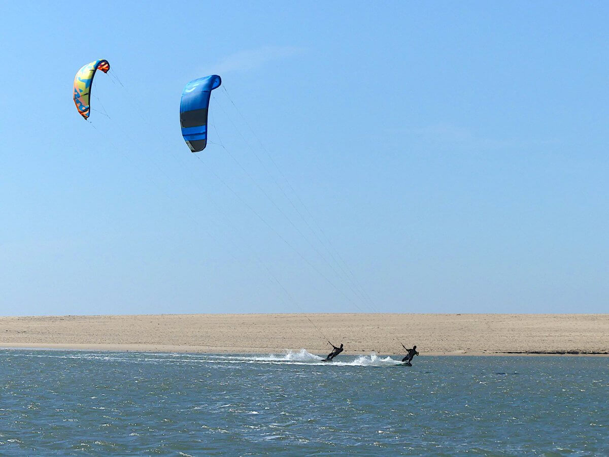 Zwei Kitesrufer flitzen über das Wasser der Lagune von Obidos, dahinter Sandstrand.