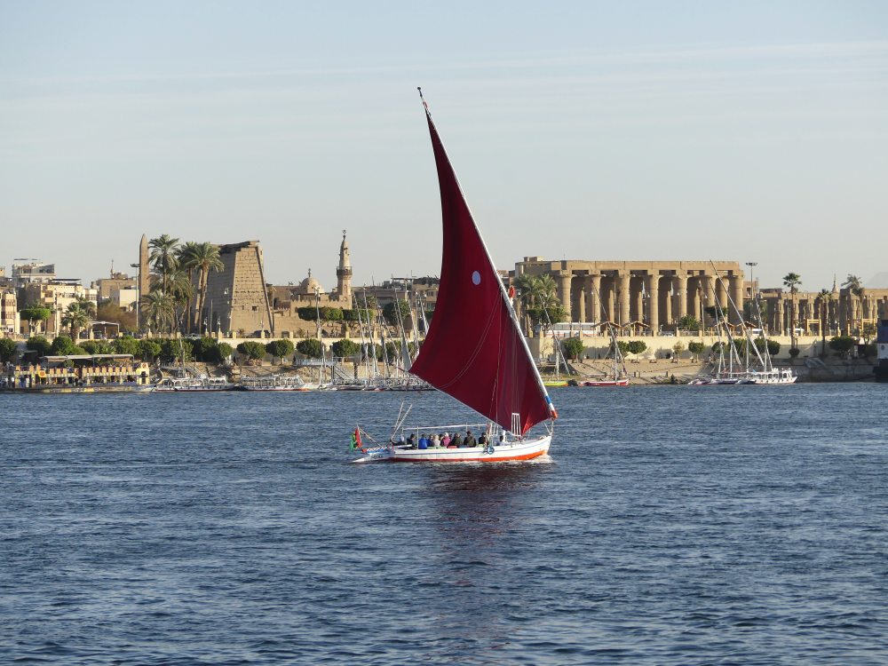 Eine Feluke mit rotem Segel fährt auf dem Nil.