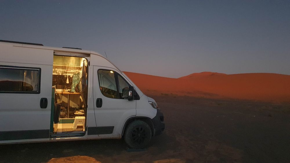 Van steht mit geöffneter Schiebetür, aus der das Licht scheint, in der Wüste am Abend.