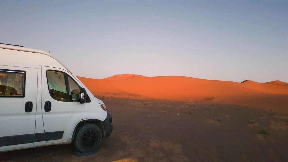 Weißes Wohnmobil steht zwischen rötlichen Dünen in der Wüste.