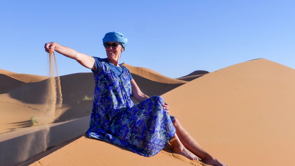 Gina sitzt in einem blauen Kleid auf dem Kamm einer Düne und lässt Sand aus ihrer Hand rieseln.