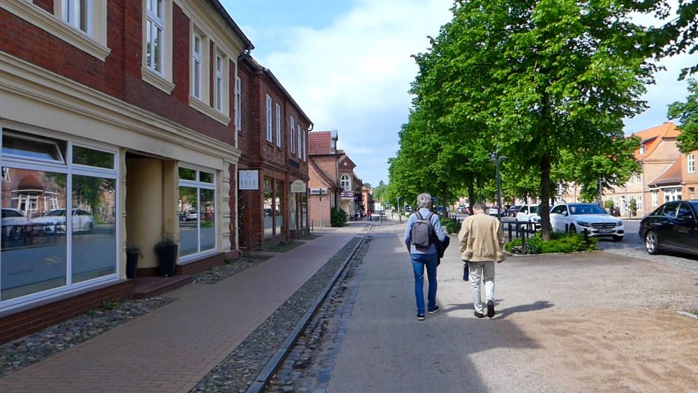 Marcus geht zusammen mit einem Mann die Straße entlang, die von historischen Häusern gesäumt ist.