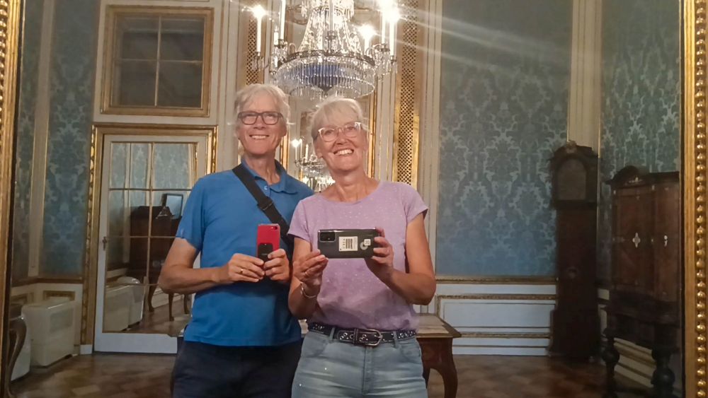 Marcus und Gina fotografieren sich vor einem großen Spiegel im Schloss, im Hintergrund ein großer Kronleuchter.