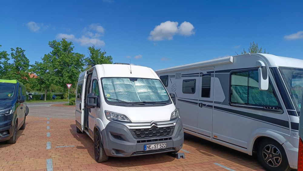 Weißer Kastenwagen steht zwischen zwei anderen Wohnmobilen auf einem Parkplatz.