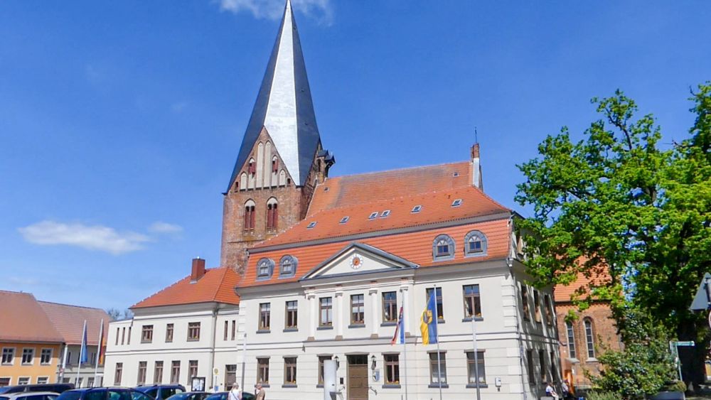 Weißes, klassizistisches Gebäude mit rotem Dach, dahinter ragt der Kirchturm empor.