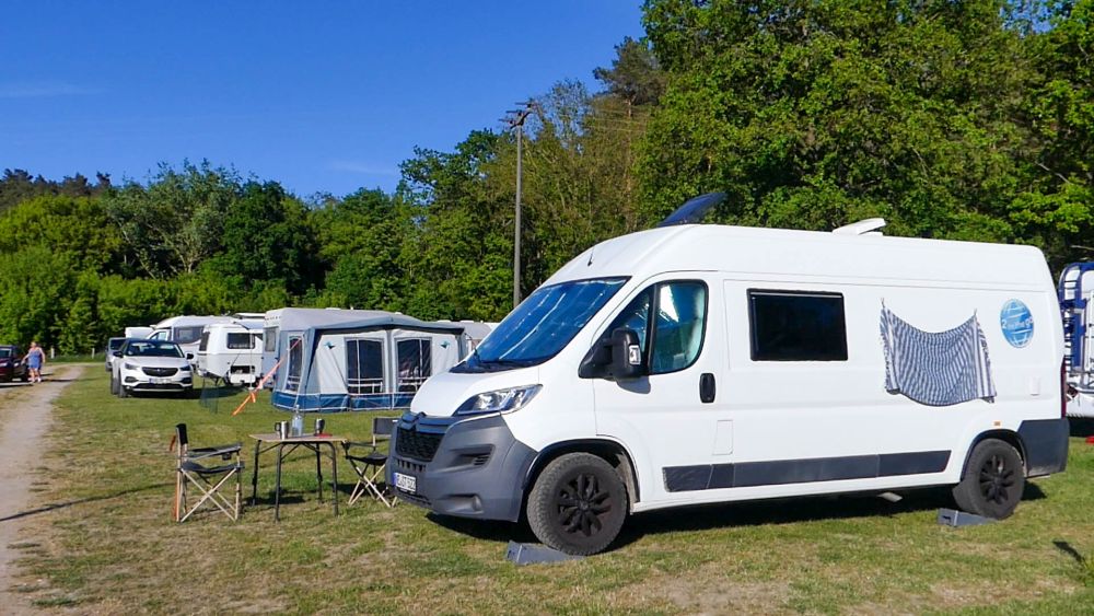 Weißer Kastenwagen steht auf dem Rasen auf dem Campingplatz. Im Hintergrund sind Wohnwagen und Wohnmobile zu sehen.