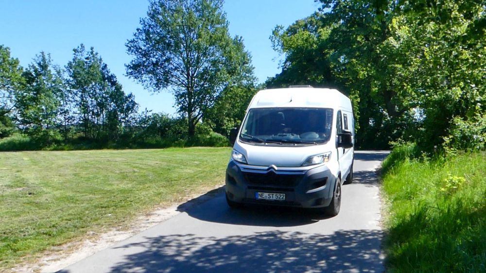 Weißer Kastenwagen faährt auf einer schmalen Straße durchs Grüne.