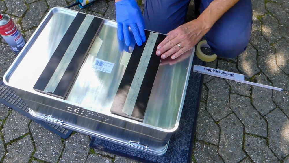 Zwei Hände, eine davon mit Schutzhandschuh, legen einen Streifen Holzplatte auf den Boden der Alubox.