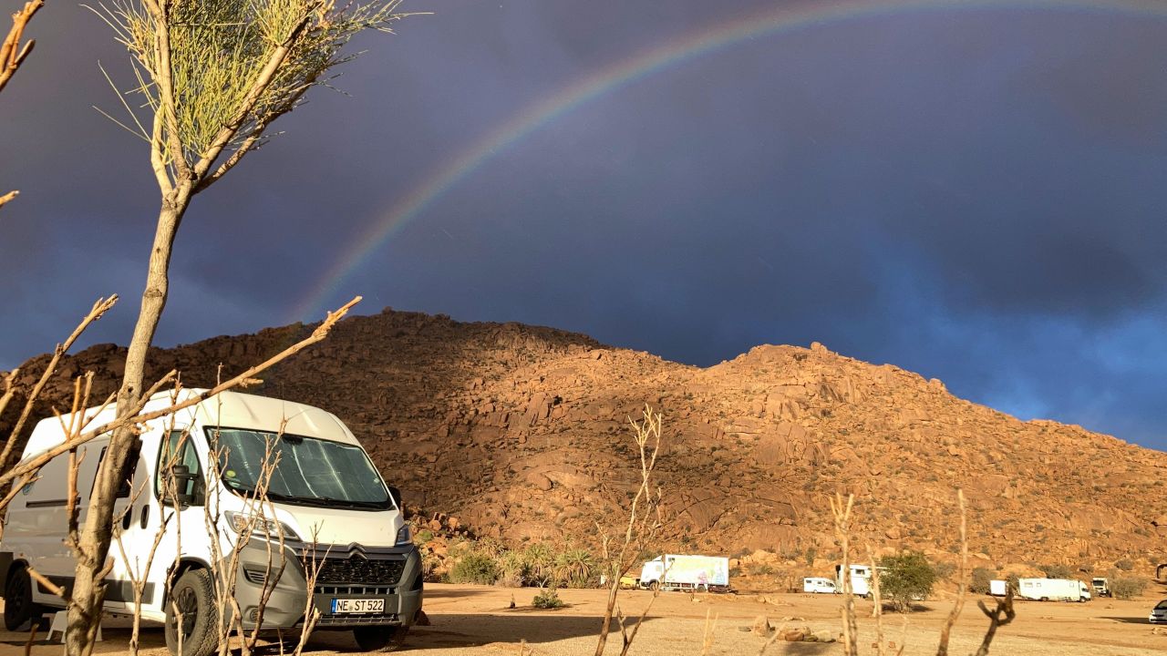 Ein weißer Van steht in einer Wüstenlandschaft mit felsigen Hügeln im Hintergrund. Ein Regenbogen wölbt sich über den bewölkten Himmel.