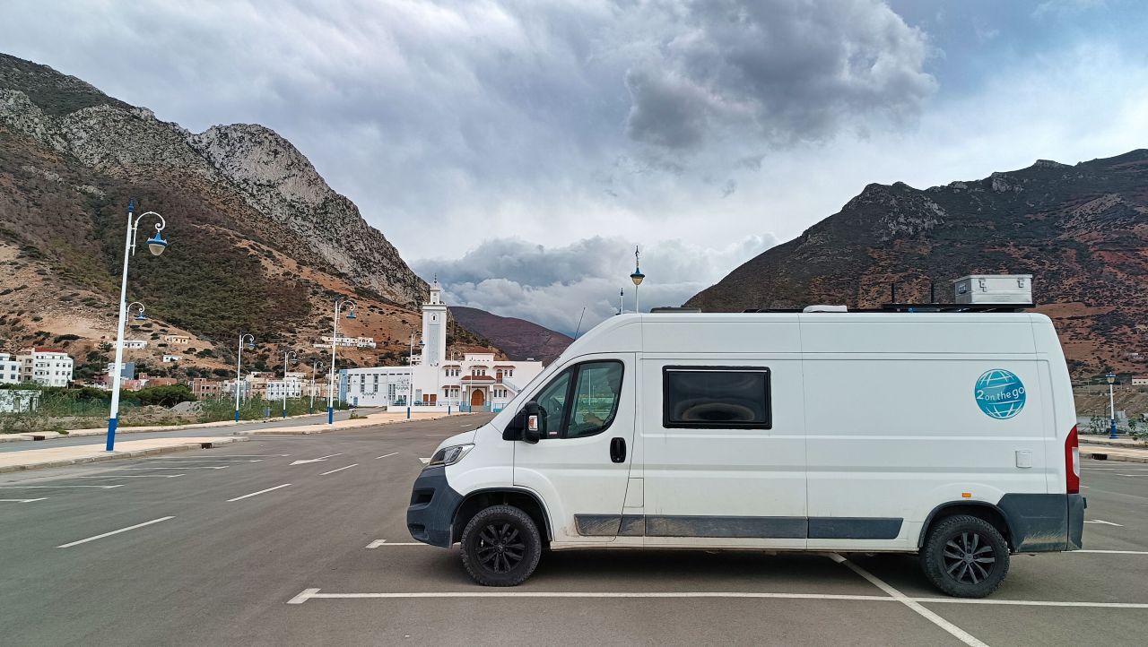 Wohnmobil steht auf einem großen Parkplatz, im Hintergrund Berge und eine Moschee.