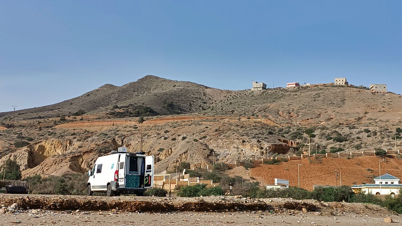 Ein Van steht mit geöffneten Hecktüren in einer wüstenartigen Landschaft mit felsigen Hügeln im Hintergrund.