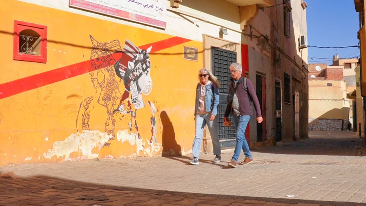 Zwei Menschen gehen unter klarem Himmel an einem farbenfrohen Wandgemälde auf einer orangefarbenen Wand in einer engen Gasse vorbei.