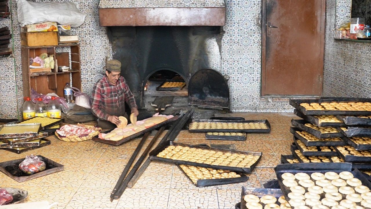 In einer traditionellen Bäckerei mit gefliesten Wänden legt eine Person Teig auf ein großes Tablett. Mehrere Tabletts mit Backwaren stehen auf dem Boden und werden in Öfen geschoben.