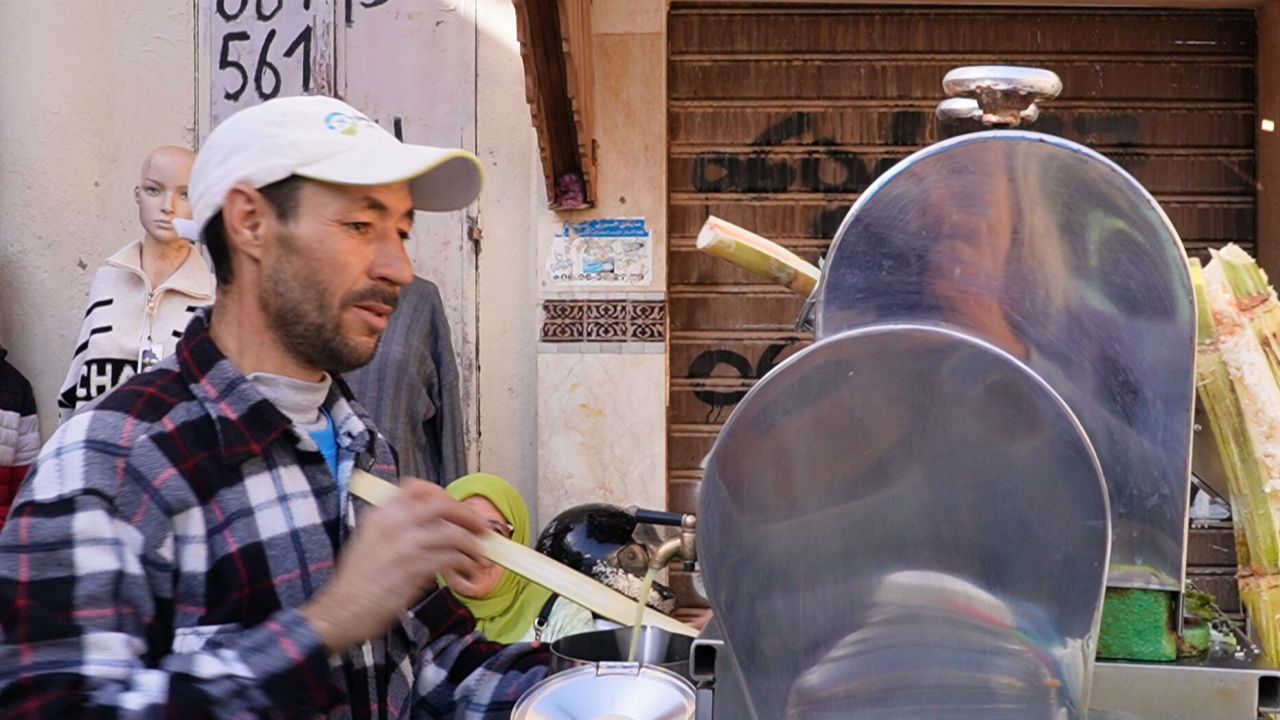 Ein Mann in kariertem Hemd und Mütze presst mit einer Maschine Saft aus Zuckerrohrstängeln. Im Hintergrund sind eine Schaufensterpuppe und ein geschlossenes Schaufenster zu sehen.